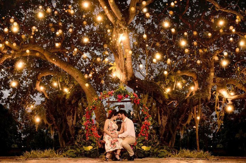 Casal sentado no balanço feito de flores, na frente da árvore principal do Arts, com diversas luzes ao redor.