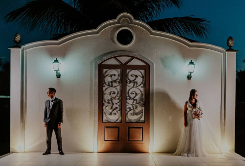 O casal de noivos em frente à nossa capela de paredes brancas. O noivo está à esquerda, olhando para a esquerda; e a noiva está à direita, olhando para a direita.